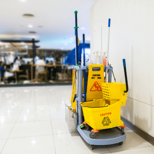 A cleaning cart with mops, a broom, a yellow mop bucket labeled "Caution," and cleaning supplies, placed in a public area.