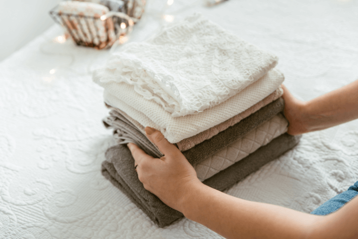 A person arranging a stack of neatly folded, soft towels in various shades of white and gray on a textured white surface.