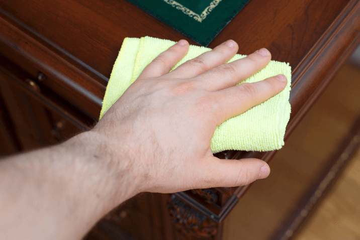 A hand holding a bright yellow cloth is cleaning the surface of a wooden table, with a green desk mat in the background.