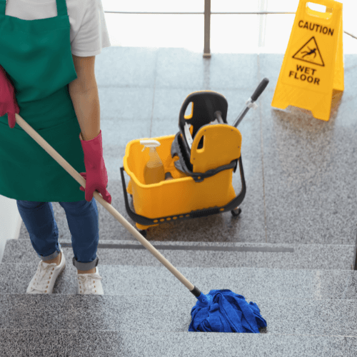 A cleaning professional mops stairs, wearing gloves and an apron, with a caution sign and a mop bucket nearby.